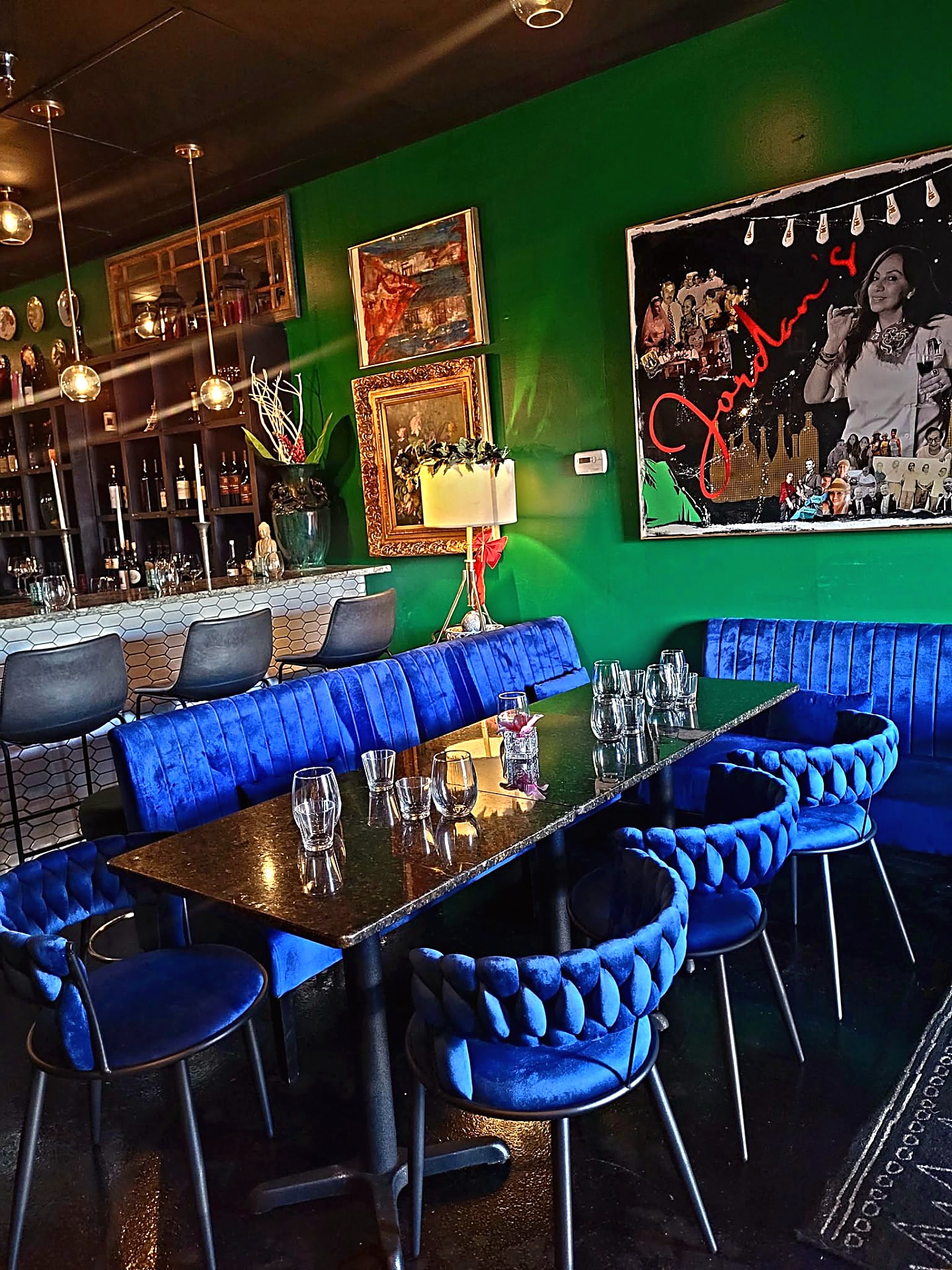 Jordan's Wine Bar interior: emerald-green wall with framed art, royal-blue velvet banquette and chairs, black stone tables set with wine glasses, and the bottle-lined bar behind